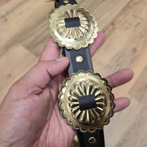 Op de afbeelding: Een zwarte leren riem met twee grote, goudkleurige concho's met geschulpte randen. De concho's hebben een centraal vierkant ontwerp en zijn verbonden door een zwarte leren riem. De riem wordt in een hand gehouden.
