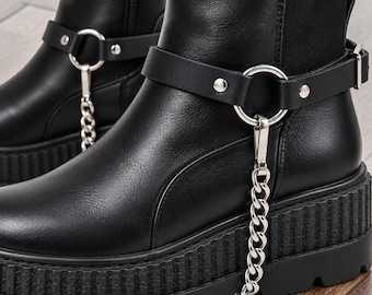 Peut inclure: Bottines noires dotées d'une épaisse semelle plateforme striée et d'une finition lisse. Chaque botte comprend une bride noire autour de la cheville fixée par un anneau en métal argenté et une chaîne argentée pendante.