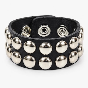 May include: A black leather cuff bracelet featuring two horizontal rows of polished silver-tone metal studs. The bracelet is secured with two silver-tone metal snap fasteners visible on the interior band, creating a circular shape.