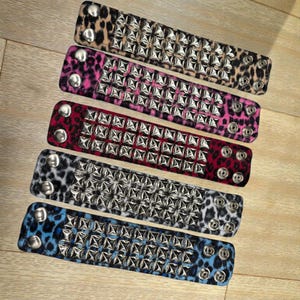 May include: Five studded wristbands in various colors and patterns. The wristbands feature a leopard print design in tan, pink, red, gray, and blue. Each band is adorned with rows of silver-colored studs and has silver snap closures.