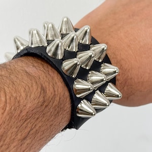 Puede incluir: Un brazalete de cuero negro con pinchos de metal plateado. El brazalete se lleva en la muñeca.