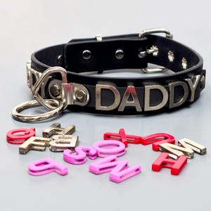 May include: A black leather collar with the word "DADDY" in silver letters. The collar has a silver metal ring and buckle. Scattered around the collar are individual letters in pink and red.