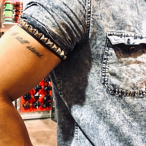 Puede incluir: Primer plano de un brazo con un tatuaje que dice "I still stand". La persona lleva una camisa vaquera con un brazalete de tachuelas en el brazo.
