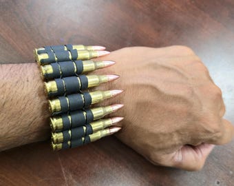 May include: A bracelet made of brass bullet casings and black connectors. The bracelet is worn on a wrist, showcasing the metallic gold and copper tones of the ammunition. The design is a unique accessory.
