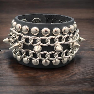 May include: A black leather cuff bracelet with silver-colored studs, chains, and cone-shaped spikes. The bracelet has a snap closure and is displayed on a dark wood surface. The design is bold and edgy.