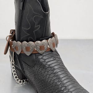 Puede incluir: Una bota de vaquero de cuero negro con una correa de tobillo decorativa de cuero marrón. La correa presenta adornos plateados en forma de corazón y una cadena plateada. La bota tiene un patrón texturizado similar a una serpiente.