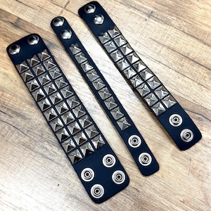 May include: Three black leather wristbands with silver pyramid studs. Each wristband has a different design, with two having multiple rows of studs and one with a single row. The wristbands have silver snap closures.