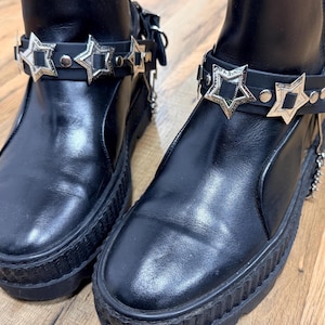 Puede incluir: Un par de botines negros con suela gruesa. Cada bota presenta una correa de cuero negro adornada con adornos plateados en forma de estrella. Las botas tienen un diseño elegante y moderno, adecuado para varios estilos de moda.