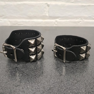 May include: Two black leather wrist cuffs with silver pyramid studs and buckle closures. Each cuff has two rows of studs. The cuffs are displayed on a dark surface.