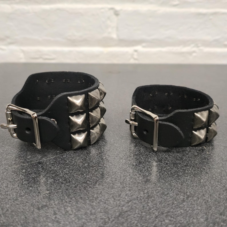 May include: Two black leather wrist cuffs with silver pyramid studs and buckle closures. Each cuff has two rows of studs. The cuffs are displayed on a dark surface.