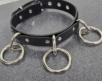 Può includere: Choker in pelle nera con anelli in metallo argentato. Il choker ha una chiusura a fibbia e più file di borchie argentate.