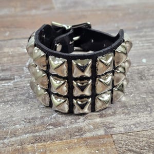 Könnte beinhalten: Schwarzes Lederarmband mit drei Reihen silberner Pyramiden-Nieten. Das Armband hat einen silbernen Schnallenverschluss. Das Armband ist für verschiedene Stile geeignet.