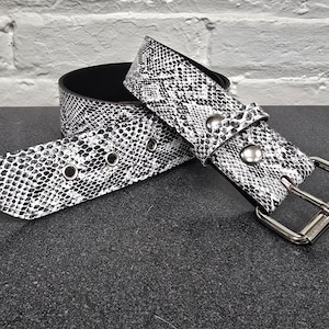 May include: A black and white snakeskin-patterned belt with a silver buckle and rivets. The belt is made of a textured material and has a black interior. The belt is unclasped and lies on a dark surface.