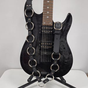 May include: Black leather guitar strap with silver rings. The strap is attached to a black electric guitar.