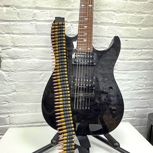 May include: A black electric guitar with a glossy finish, featuring two black humbucker pickups labelled Custom Alnico, a dark wood fretboard with diamond-shaped inlays, and a black ammunition belt strap draped vertically across the guitar body.