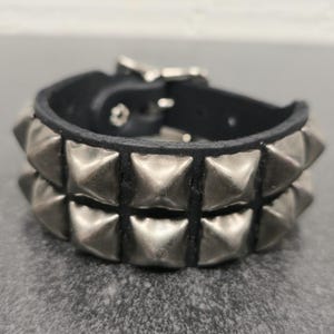 May include: A black leather bracelet with two rows of silver-colored pyramid studs. The bracelet has a silver buckle closure. The design is edgy and suitable for a rock and roll or punk aesthetic.