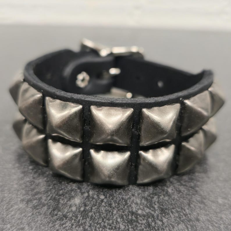 May include: A black leather bracelet with two rows of silver-colored pyramid studs. The bracelet has a silver buckle closure. The design is edgy and suitable for a rock and roll or punk aesthetic.