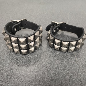 May include: Two black leather wristbands with silver pyramid studs. Each wristband has three rows of studs and a silver buckle closure. The wristbands are displayed on a gray surface.