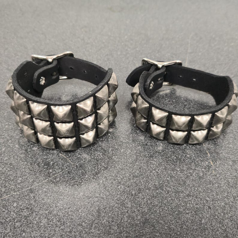 May include: Two black leather wristbands with silver pyramid studs. Each wristband has three rows of studs and a silver buckle closure. The wristbands are displayed on a gray surface.