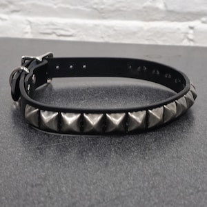 May include: Black leather collar with silver pyramid studs. The collar has a buckle closure and a metal loop. The studs are evenly spaced along the collar's length, creating a bold, edgy look. The collar is designed for pets.