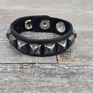 May include: A black leather bracelet with silver pyramid-shaped studs. The bracelet has a snap closure.