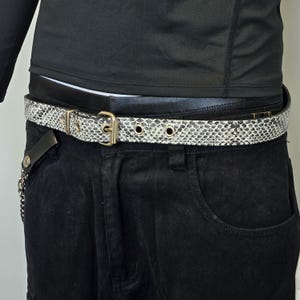 May include: A black leather belt with a snakeskin pattern and a silver buckle. The belt is worn around the waist of a person wearing black pants.
