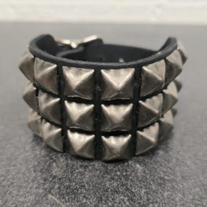 May include: A black leather bracelet with three rows of silver-colored pyramid studs. The bracelet has a buckle closure and is designed to be worn around the wrist. The studs are evenly spaced and give the bracelet a bold, edgy look.