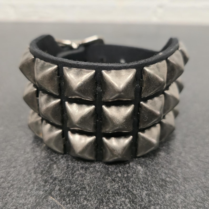 May include: A black leather bracelet with three rows of silver-colored pyramid studs. The bracelet has a buckle closure and is designed to be worn around the wrist. The studs are evenly spaced and give the bracelet a bold, edgy look.