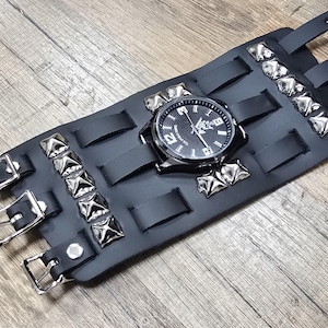 May include: A black leather wristband watch with a silver-tone watch face and silver-tone pyramid studs. The watch has a black leather strap and buckle closure. The watch face has white numerals and hands.