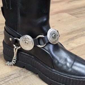 Puede incluir: Bota de cuero negro con una correa plateada que presenta dos medallones circulares decorativos. La correa está conectada por un anillo plateado y una cadena. La bota tiene una suela gruesa y negra.