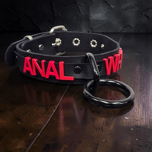 Pode incluir: Uma coleira preta com letras vermelhas que formam a palavra ANAL e parte da palavra WH. A coleira inclui uma fivela prateada, ilhós de metal e um grande anel preto fixado na parte frontal, com um acabamento liso.