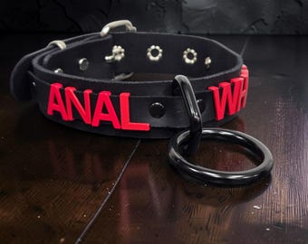 May include: A black collar featuring red lettering that spells out the word ANAL and part of the word WH. The collar includes a silver-toned buckle, metal eyelets, and a large black O-ring attached to the front, constructed with a smooth finish.