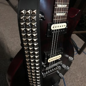 May include: A black leather guitar strap with silver pyramid studs, draped over a dark red electric guitar. The guitar has silver tuning pegs and a white pickup.
