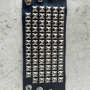 May include: A black leather bracelet with silver pyramid-shaped studs. The bracelet has five silver round studs on the top edge.