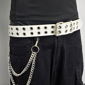 May include: White belt with multiple eyelets and a silver buckle. A silver chain hangs from the belt, attached to black pants. The belt is worn with a black shirt.