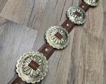 May include: A brown leather belt featuring a series of evenly spaced, circular silver-toned metal conchos. Each concho has a scalloped edge and a central rectangular brown leather inlay. Small silver-toned metal rivets secure the conchos to the belt.