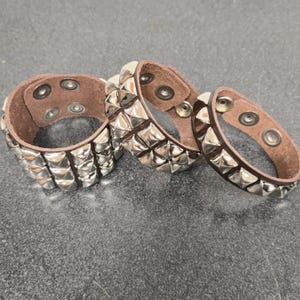 Puede incluir: Tres pulseras de cuero marrón con filas de tachuelas plateadas en forma de pirámide. Cada pulsera tiene un cierre de botón a presión. Las pulseras se muestran sobre una superficie gris oscuro.