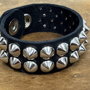 Könnte beinhalten: Ein schwarzes Lederarmband mit zwei Reihen silberner pyramidenförmiger Nieten.
