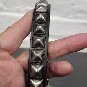 May include: Black leather bracelet with silver square studs. The bracelet is a close-up shot, showcasing the texture of the leather and the metallic sheen of the studs. The design is a classic punk or rock-inspired accessory.