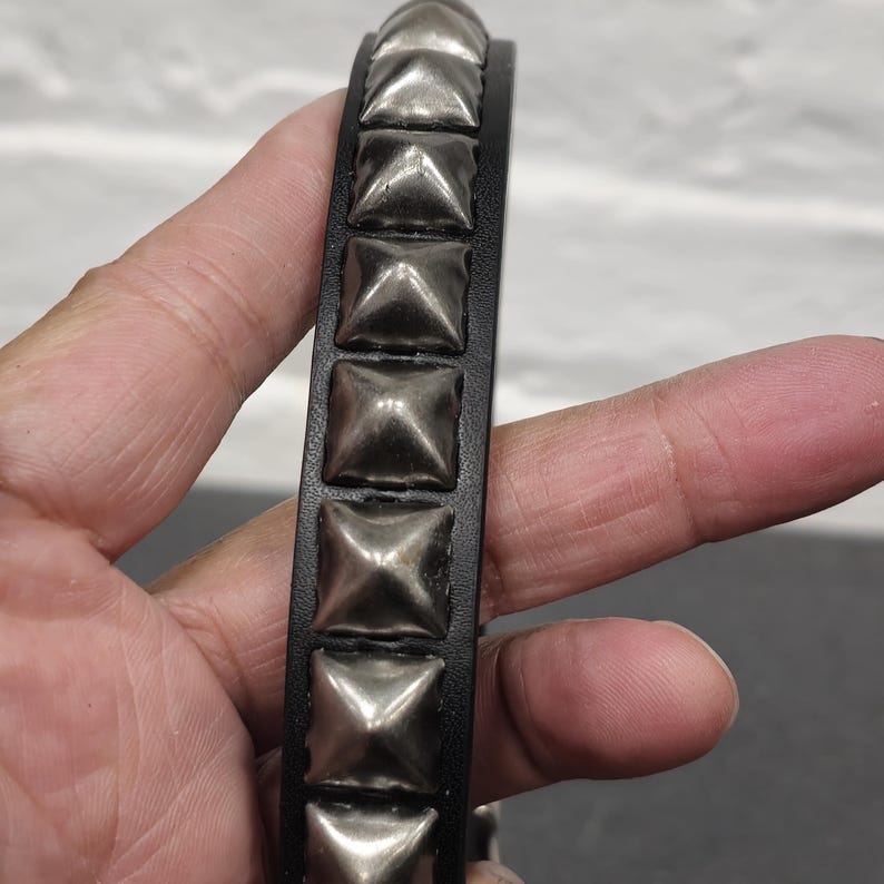 May include: Black leather bracelet with silver square studs. The bracelet is a close-up shot, showcasing the texture of the leather and the metallic sheen of the studs. The design is a classic punk or rock-inspired accessory.