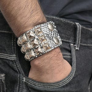 May include: A silver studded bracelet with a snakeskin pattern is worn on a wrist. The bracelet features rows of large, shiny silver studs and is secured with two silver snaps. The person is wearing dark denim jeans.