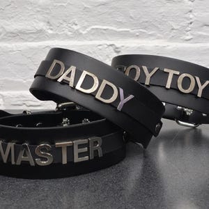 Può includere: Tre collari in pelle nera con lettere in metallo argentato. I collari recitano "MASTER", "DADDY" e "BOY TOY".