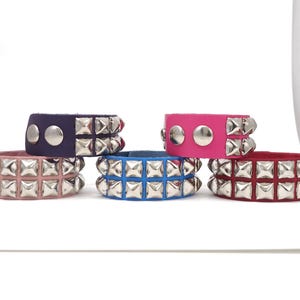 May include: A collection of colorful studded leather bracelets. The bracelets are in various colors, including pink, blue, purple, and red. Each bracelet features silver-colored studs and a silver snap closure. These bracelets are a bold fashion accessory.
