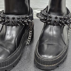 Puede incluir: Un par de botas de cuero negro con suelas gruesas y correas ajustables en el tobillo. Cada correa está adornada con dos filas de púas negras afiladas e incluye un detalle de cadena de metal plateado sujeto al lateral de la bota.