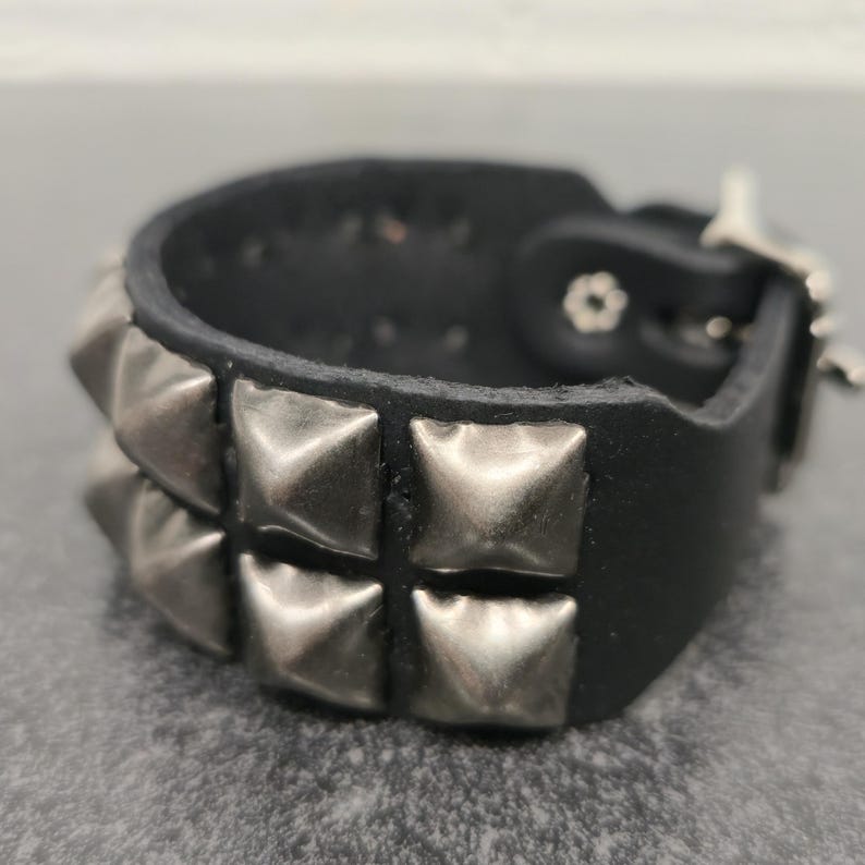 May include: Black leather bracelet with two rows of silver-colored pyramid studs. The bracelet has a buckle closure and is designed to be worn around the wrist. The style is reminiscent of punk rock fashion.