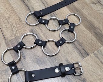 May include: A black leather belt with silver metal rings. The belt has a buckle and is adjustable.