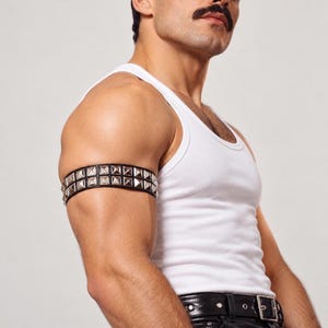 May include: A black leather armband featuring two rows of silver-toned pyramid studs. The band is worn on the upper arm, complementing a white ribbed tank top and a black leather belt with a silver-toned metal buckle.