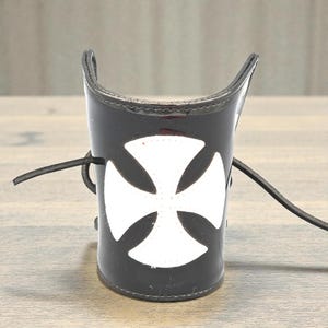 May include: Black leather arm cuff with a white Maltese cross. The cuff has a glossy finish and a lace-up closure. The design is a striking accessory.