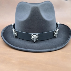 May include: A dark gray fedora hat with a black band adorned with silver fox head accents. The hat has a classic teardrop crown and a wide brim. The hat is made of a felt-like material.