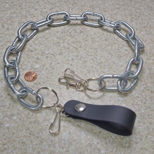 Könnte beinhalten: Eine silberne Metallkette mit einem Schlüsselring, einem Verschluss und einer dunkelgrauen Lederschlaufe. Ein Cent-Stück liegt neben der Kette zum Größenvergleich. Die Kette besteht aus großen, ovalen Gliedern.